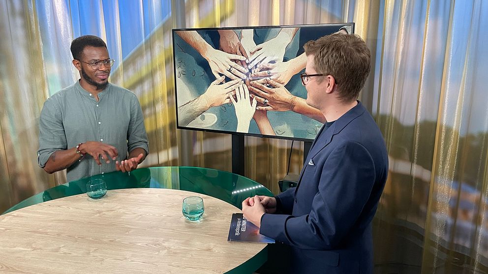 Romel Belcher och Magnus Bergevin står vid ett runt bord i en studio. I bakgrunden visas en tv-skärm en illustrerad bild med flera händer som symboliserar samarbete och gemenskap. På bordet finns två vattenglas.