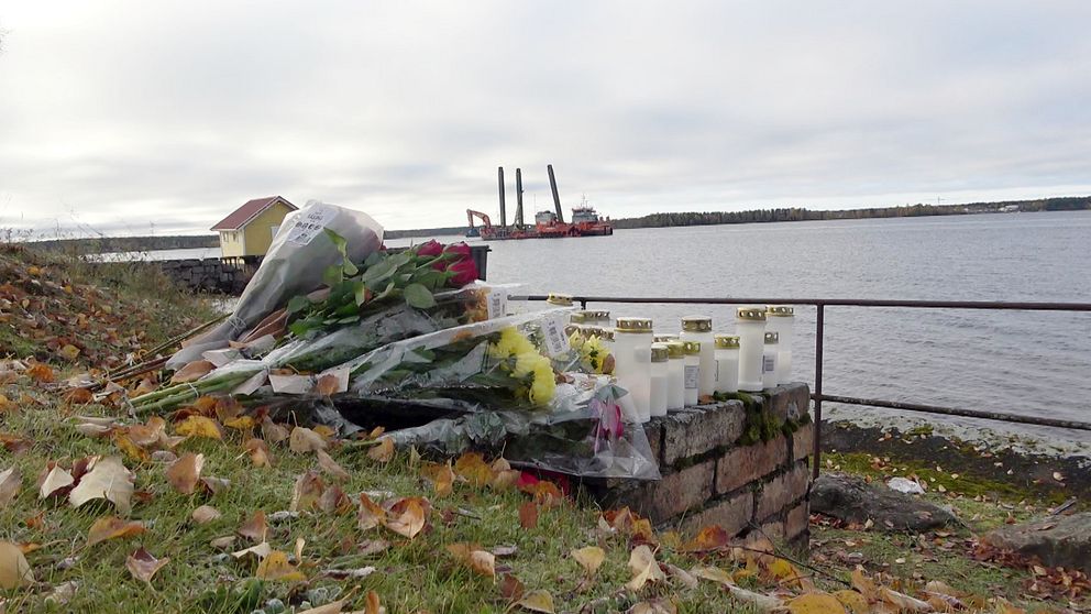 Blommor och tända ljus ligger på en mur vid vattnet, omgivna av fallna löv. I bakgrunden syns en muddringsbåt. Himlen är grå och molnig.