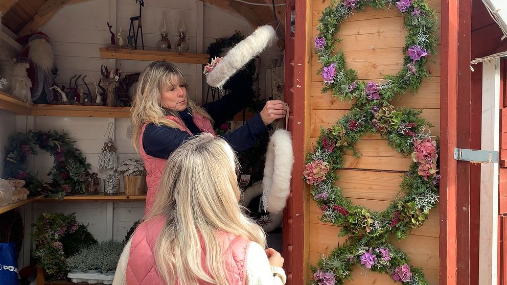 Två personer arbetar i en marknadsbod där det säljs blomsterdekorationer.  Det finns flera dekorationer, inklusive kransar, i bakgrunden, vilket skapar en mysig och kreativ atmosfär.