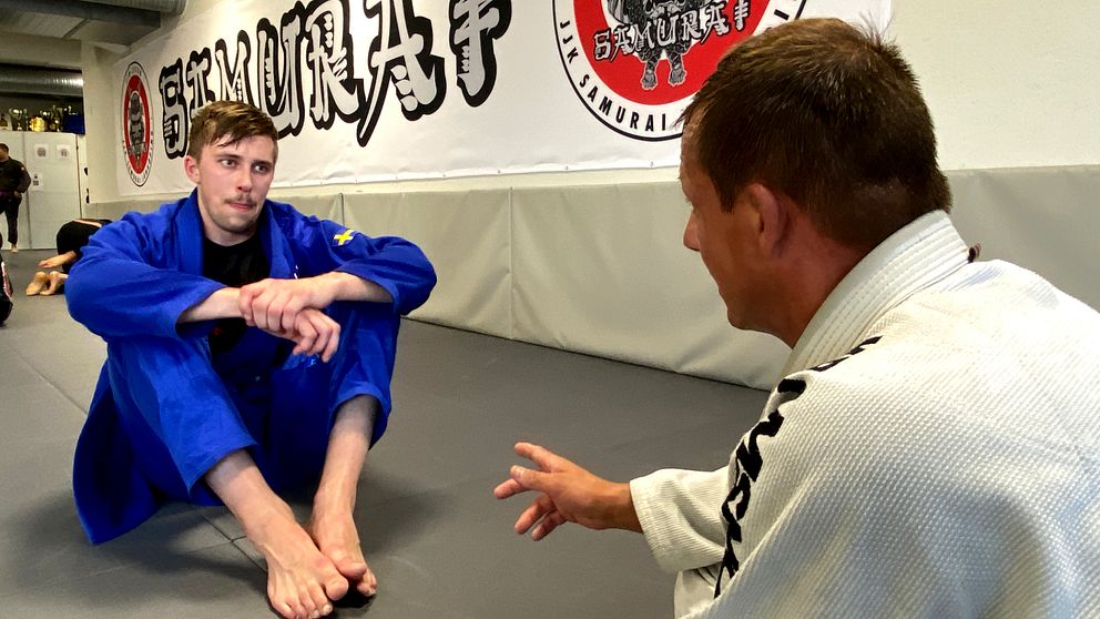 man i blå kampsportsdräkt får instruktioner av annan man på golvet i en kamsportslokal