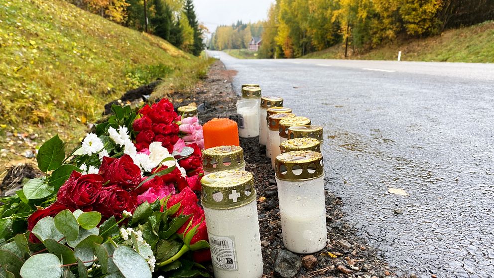 Blommor i röd och vit färg ligger på marken vid vägkanten, omgivna av flera ljus med guldkant som är tända. I bakgrunden syns en stilla väg med träd som har höstfärger. Bilden förmedlar en känsla av minne och sorg.