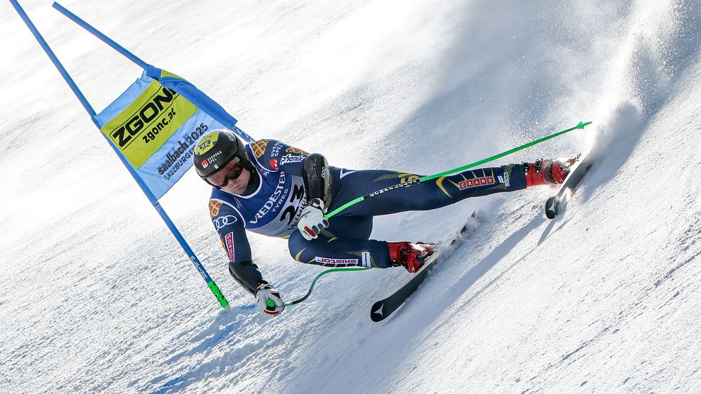 En alpin skidåkare i full fart svänger genom en blå port under en slalomtävling på en snötäckt backe. Skidåkaren bär en mörk dräkt med olika sponsormärken och är fokuserad på att manövrera genom banan.
