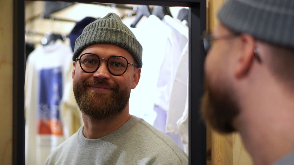 En person i en grå tröja och en grå beanie står framför en spegel i en klädaffär. Bakgrunden visar klädesplagg som hänger på galgar, vilket skapar en butiksmiljö.