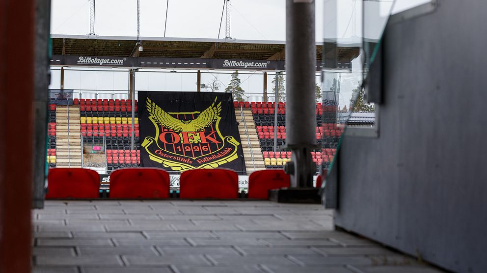 Stor svart banner med Östersunds klubbemblem hänger framför tomma läktare på en fotbollsarena, med rader av röda och gula stolar synliga. Bilden fångar en vy från en ingång till arenan, vilket ger en känsla av förväntan inför en match.