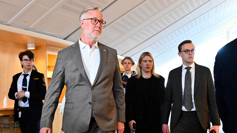 En grupp personer i kostym och kavaj går i en korridor, ser allvarliga ut.
