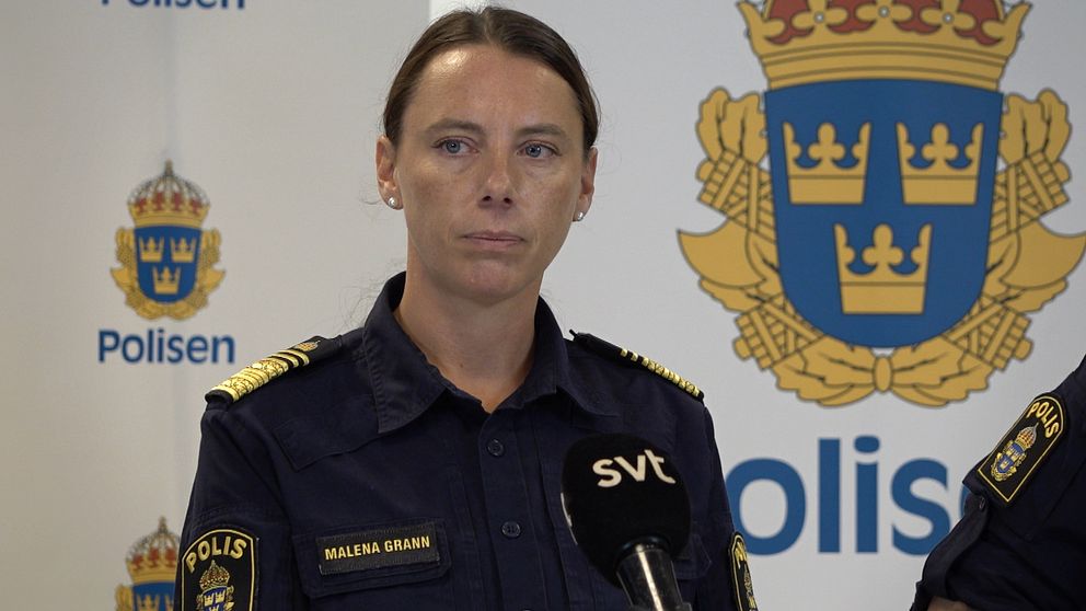 Kvinnlig polis står i uniform framför vit vägg med polisens logotyper på.
