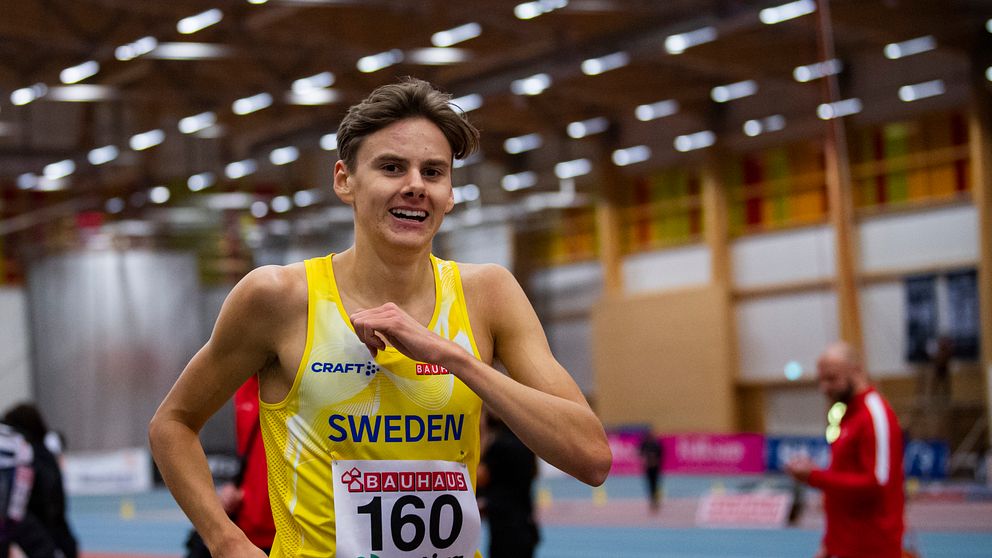 En löpare i tävlingskläder med texten ”SWEDEN” och nummerlappen 160 springer inomhus på en löparbana. I bakgrunden syns tränare och andra deltagare i en stor hall med träinredning och neonljus.
