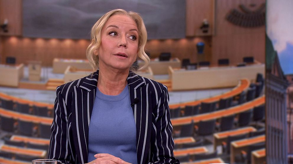 Kvinna i kavaj framför en bakgrund av riksdagens plenisal.