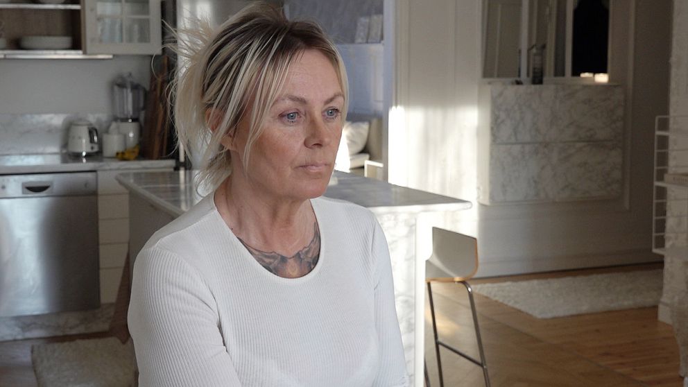 Kvinna i vit långärmad tröja sitter i ett modernt kök med vita marmordetaljer och trädetaljer i bakgrunden.