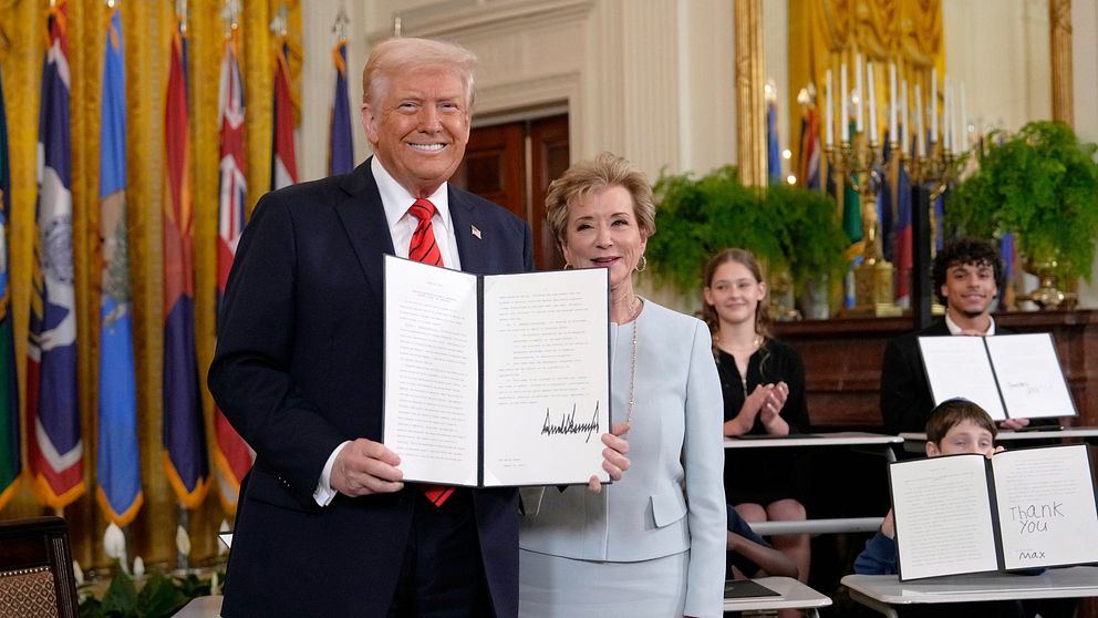 Dobald Trump håller upp ett dokument. I bakgrunden ser man flaggor och applåderande personer.