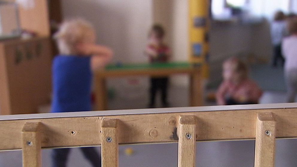 Interiör av en förskola med små barn som leker i bakgrunden och en träbarriär i förgrunden. Barnen är upptagna med olika aktiviteter i ett rymligt rum.