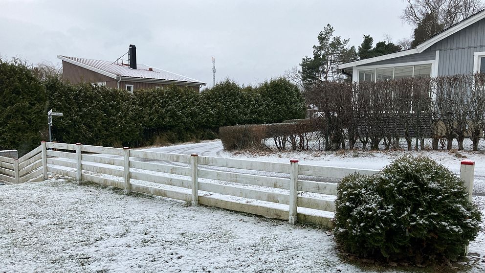 Snötäckt trädgård med vita staket och ett grått hus i bakgrunden.