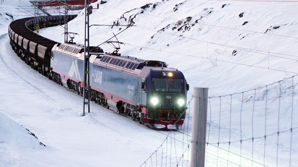 Ett av LKAB:s tåg med flera malmvagnar färdas längs en snöigt järnvägslinje. Omgivningen består av snöklädda berg och en elektrisk stolpe nära spåret.