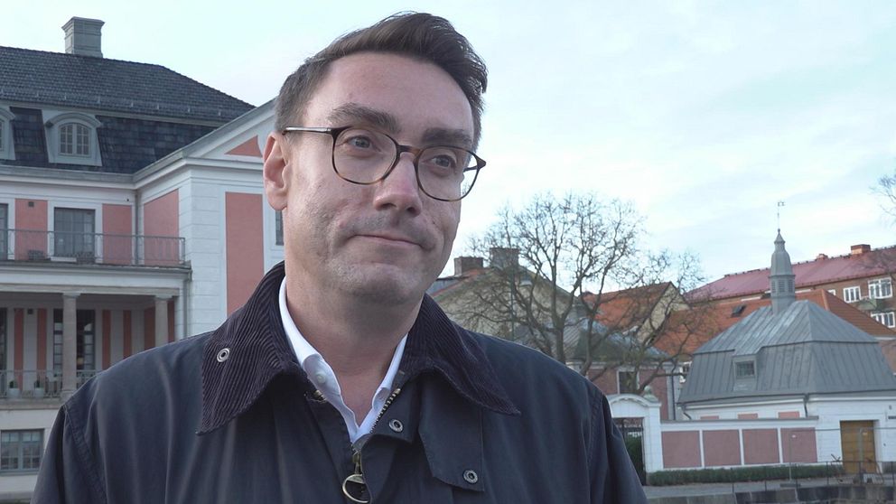 En person står framför en historisk byggnad med vit och rosa fasad. I bakgrunden syns träd och andra hus, vilket skapar en stadsbild. Personen bär en mörk jacka och en vit skjorta. Himmelns nyanser är ljusa med några moln.