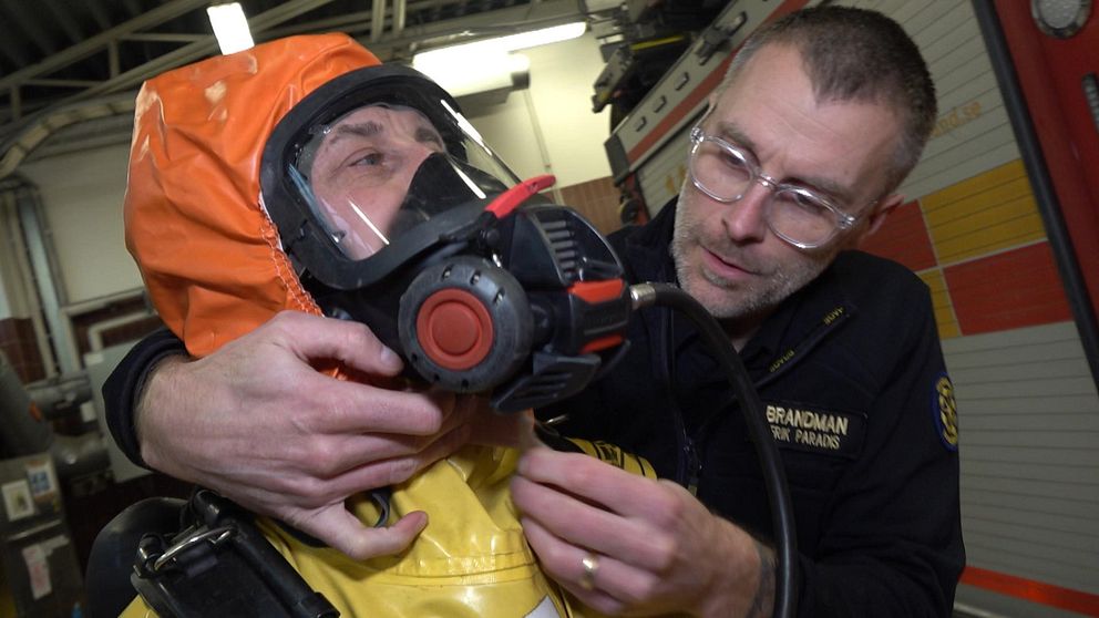 Brandmän hjälper varandra med att sätta på sig utrustning för att kemdyka. En man hjälper en anna med att sätta på dig en orange galonhätta som ska skydda mot giftiga kemikalier. Mannen med hätta har en stor ansiktsktsmask på sig.