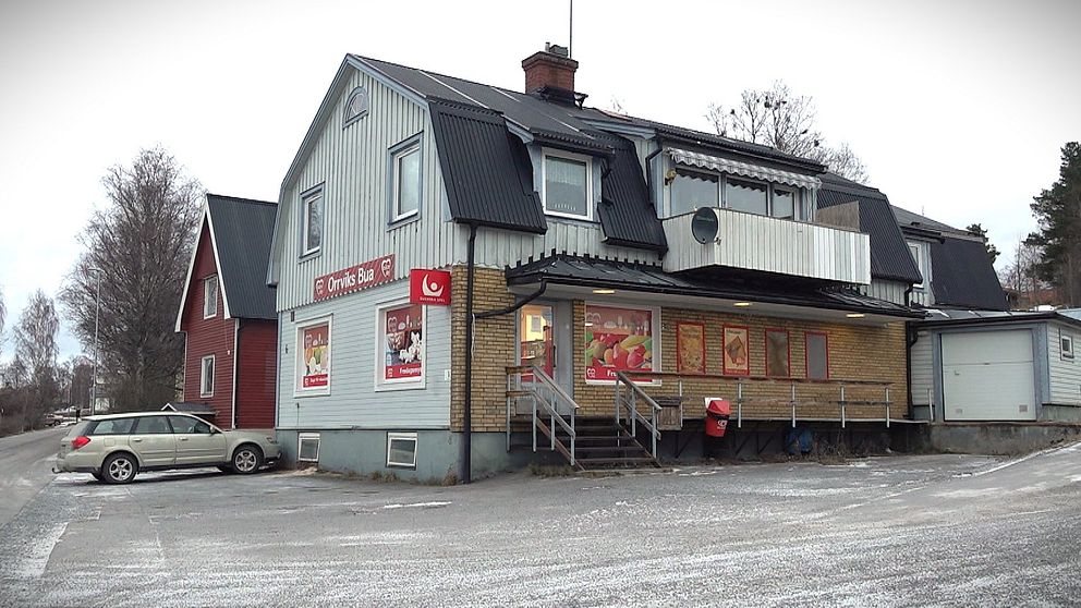 Lanthandeln Orrviksbua med en kombination av trä- och tegelkonstruktion. Framsidan har stora fönster med affischer och några trappor som leder upp till ingången. Parkerade bilar finns i närheten, och det är vinterväder med snö på marken. Runt omkring syns flera träd och andra byggnader, vilket skapar en lugn atmosfär.