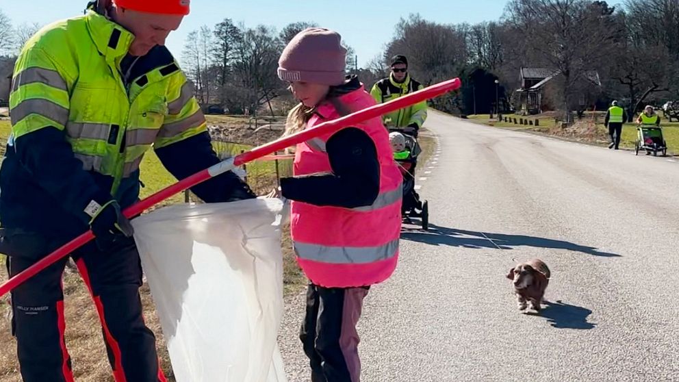 Två personer städar vägkant, en liten hund står på vägen.