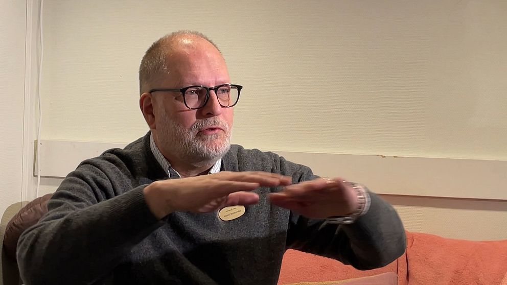 En person sitter i en soffa och gestikulerar med händerna, iklädd en grå tröja och en namnbricka. Bakgrunden är en ljus vägg med en horisontell list.