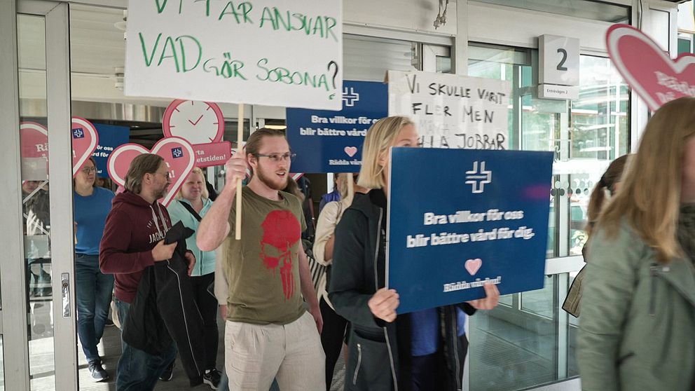 Vårdpersonal strejkar med plakat vid entrén till ett sjukhus.