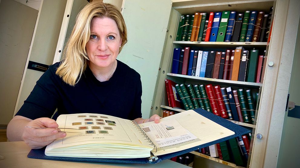 Anna Johnsson Winwvist på auktionshuset Postiljonen bläddrar i bok med frimärken vid bokhylla.