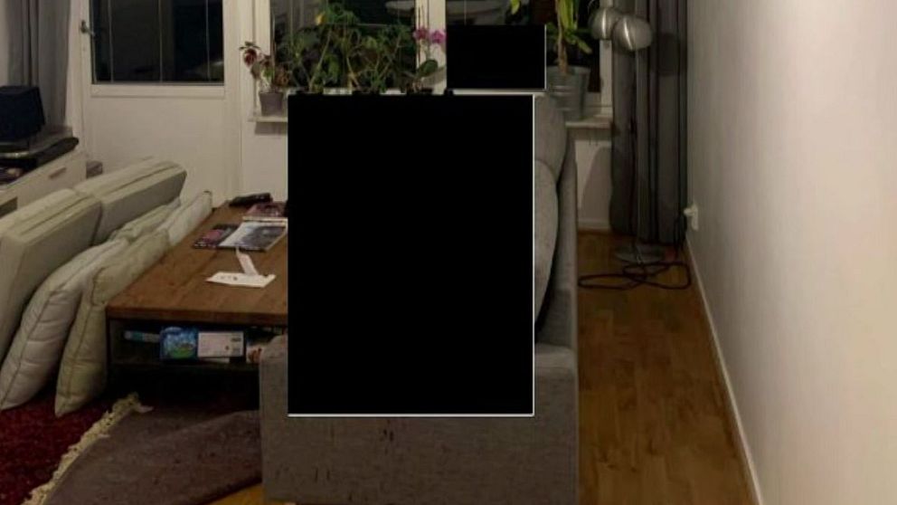 Vardagsrum med en hörnsoffa i grått och ett soffbord i trä. I bakgrunden syns stora fönster som släpper in ljus, samt växter i fönstret. En TV står på en vit mediabänk. Golvet är av trä, och det finns en röd matta delvis synlig. Över soffan är en stor svart ruta.