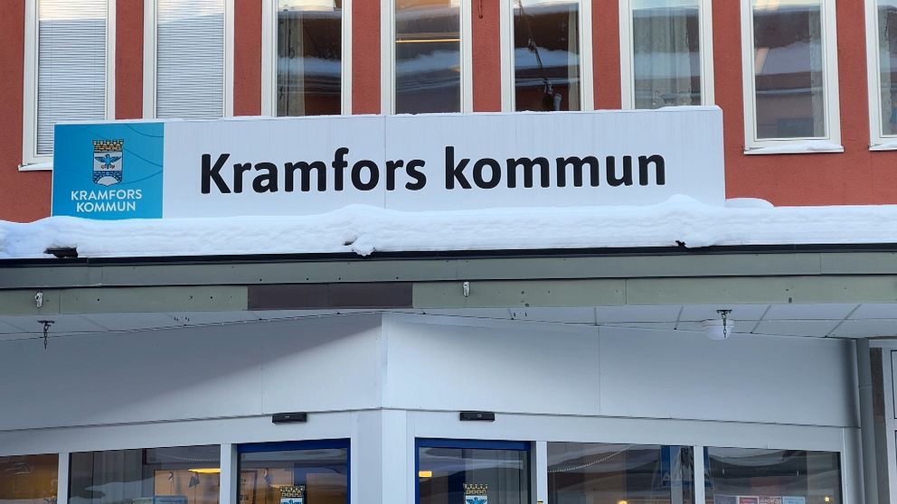 Skylt med texten ”Kramfors kommun” på en byggnad, med snö på taket och fönster i bakgrunden.