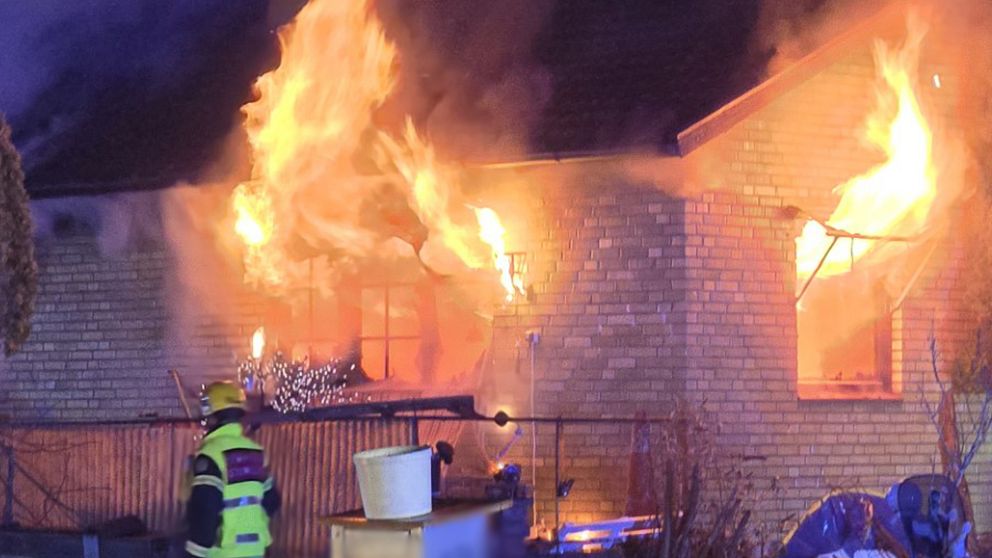 Ett tegelhus täckt av lågor under en brand, med en brandman i gul skyddsutrustning i förgrunden som verkar vara på väg mot eldsvådan. Elden ryker intensivt från husets fönster.
