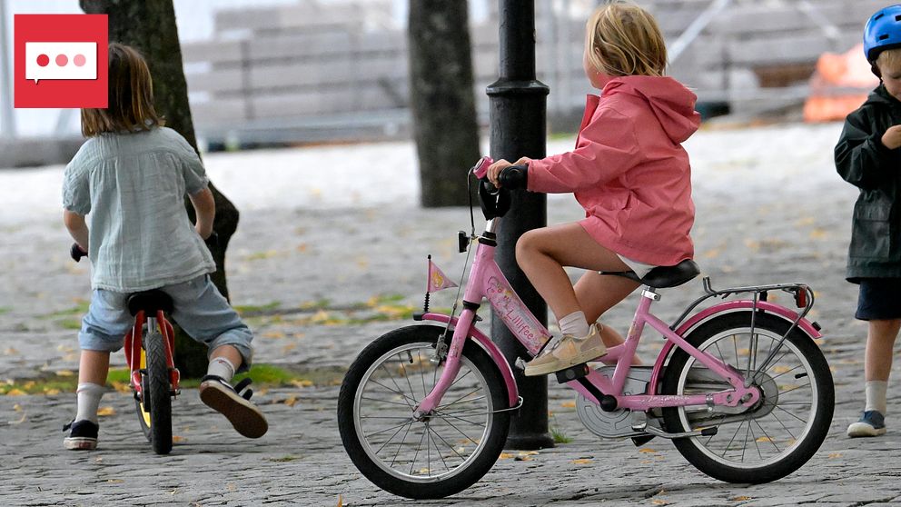 Två barn cyklar på en grusväg; ett i rosa jacka och ett med långt hår. Ett barn står bredvid.