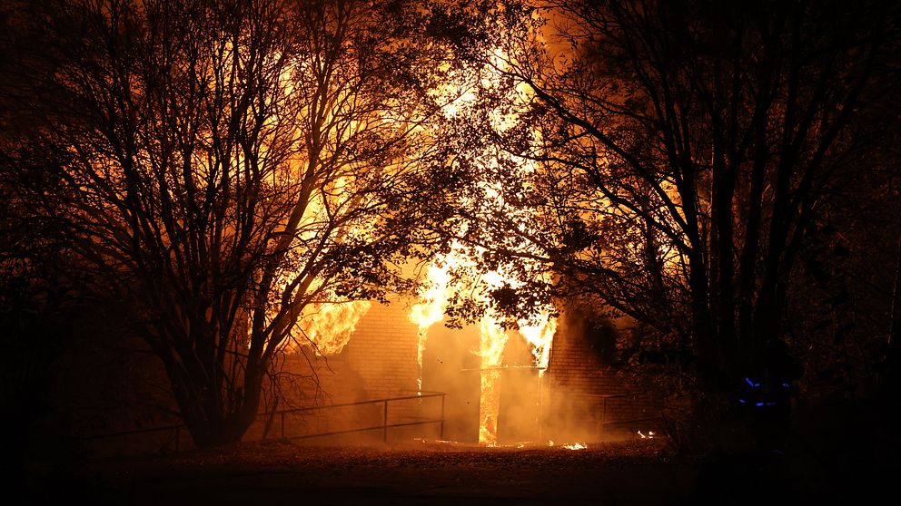 Ett hus omgärdat av stora lågor som sprider sig kraftigt, med träd som skymmer en del av byggnaden. Scenen präglas av en intensiv brand som lyser upp omgivningen i orangeröda och gula nyanser. Rök kan också ses i luften runt omkring.