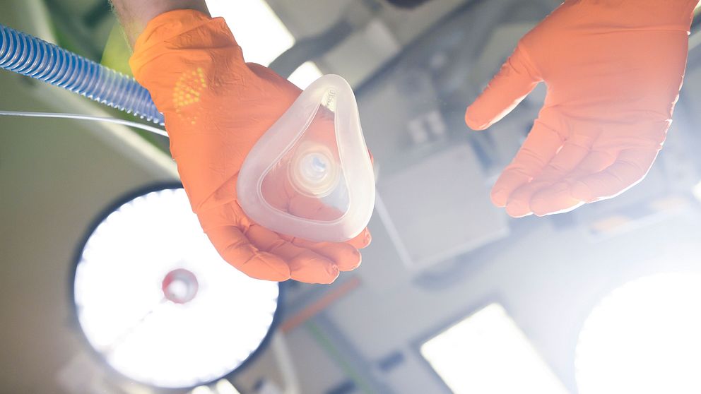 En person med orangea handskar håller en anatomisk respiratormask i handen, medan de ser ner mot den. I bakgrunden syns svagt upplysta kirurgiska lampor och en del av en medicinsk miljö, vilket tyder på att bilden är tagen i en operationssal eller liknande.
