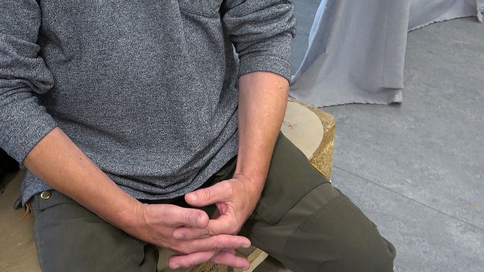 En bild på en person som sitter på en stol med händerna i knät. Bilden är beskuren så man bara ser från bröstet och nedåt.