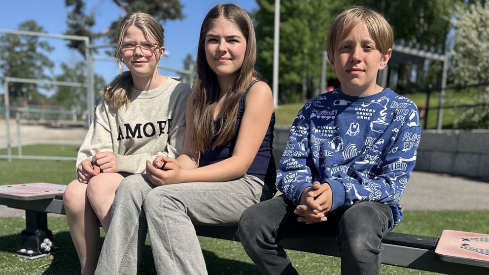 Tre elever, två tjejer och en kille som går i årskurs fem sitter på en bänk utomhus på skolgården.