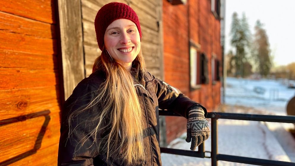 En person står lutad mot ett räcke framför en gammal träbyggnad med rödbrun fasad. Personen har på sig en mörk jacka och en röd mössa, och deras lång hår hänger ner över axlarna. I bakgrunden syns snö och träd, vilket tyder på kallt väder. Solens strålar belyser scenen och skapar en varm atmosfär.