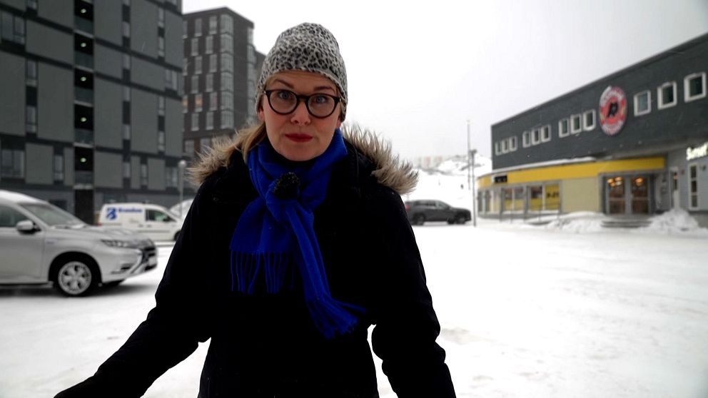 Person står utomhus på en snötäckt gata, med moderna byggnader i bakgrunden.