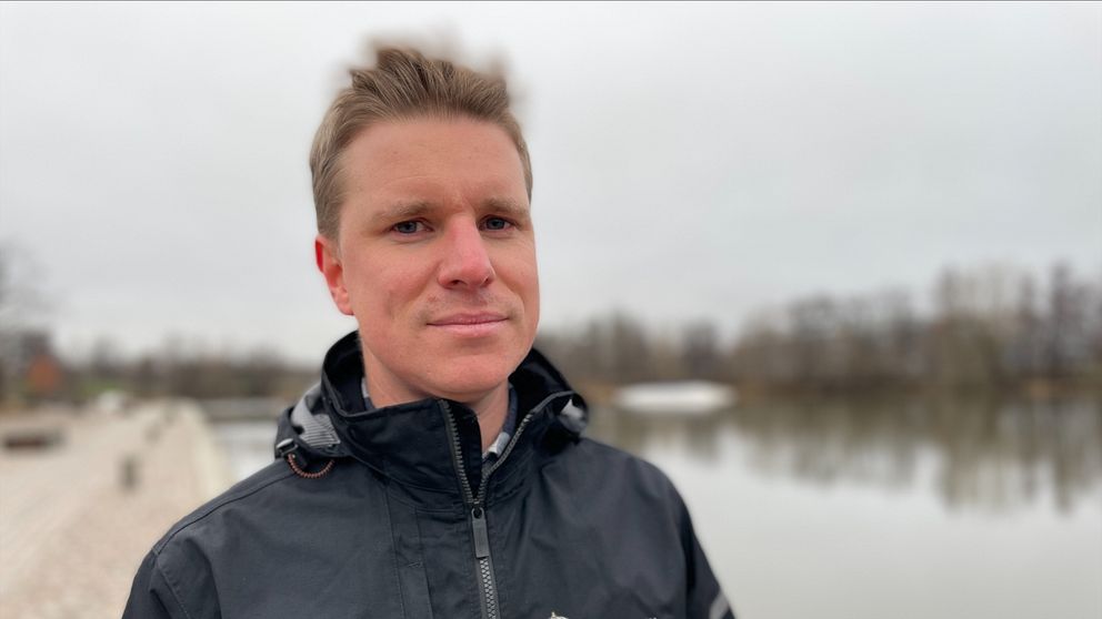 En person står vid vattnet med en mörk jacka som har en logotyp på bröstet. I bakgrunden syns en stilla sjö och träd längs strandlinjen under en grå himmel.