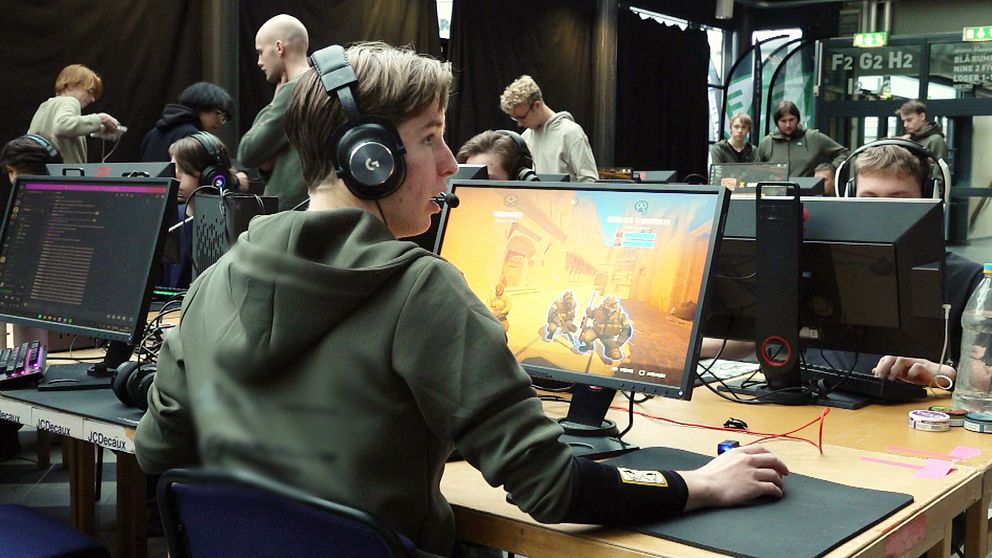En grupp personer i en spelturnering, där fokus ligger på en spelare som sitter vid en dator med hörlurar. Skärmen visar en actionsekvens från ett videospel. Omkring finns andra deltagare och en publik som observerar evenemanget. Atmosfären är livlig och fokuserad på spelande.