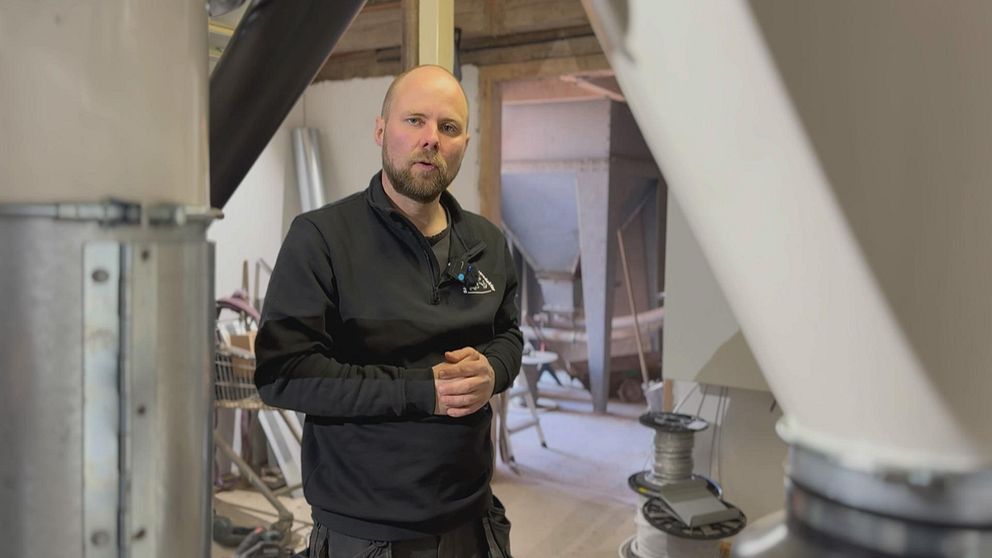 En person med svarta arbetskläder står inomhus bland stora industrimaskiner och rör.