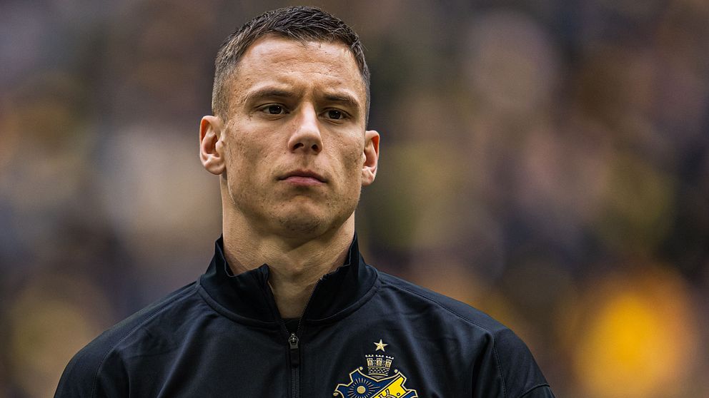 AIK-spelare med mörkt jacka och klubbmärke synligt, suddig publik i bakgrunden.