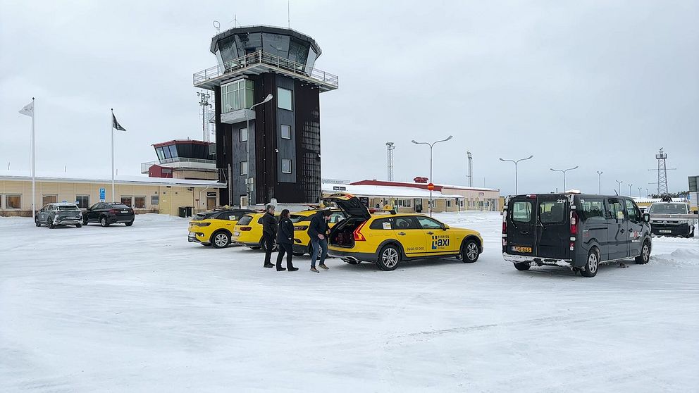 En vinterbild av Örnsköldsviks flygplats med snö på marken och flygledartornet i bakgrunden. Flera gula taxibilar står parkerade i förgrunden där några personer lastar bagage ur en taxi.