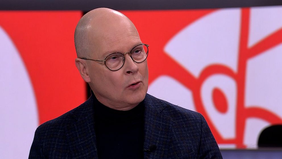 Mats Knutson i kostym står framför en färgglad bakgrund med röda och vita mönster, vilket indikerar en tv-studio eller liknande miljö. Personen verkar vara involverad i en diskussion eller intervju.