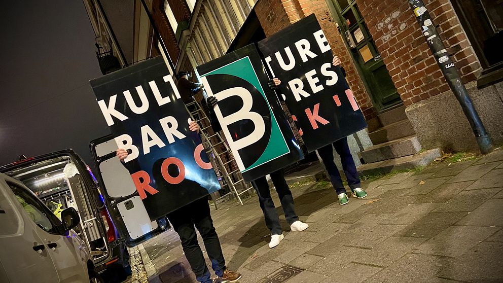 Tre personer håller upp stora skyltar som visar texten ”KULTUR”, ”BAR”, ”ROCK” och ”B” i olika färger och storlekar. Bakgrunden är en stadsmiljö på kvällen, med låg belysning och några byggnader synliga.