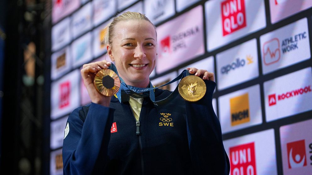 En person i en mörk blå jacka håller upp två guldmedaljer framför en vägg med sponsormärken och logotyper, vilket tyder på en framstående prestation inom sport.