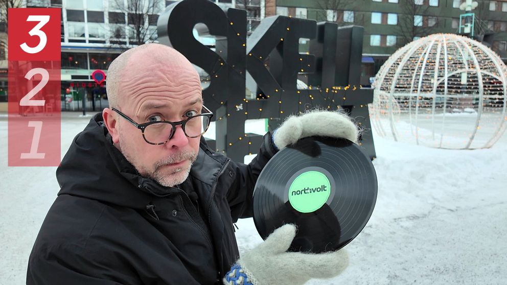 Reporter Patrik Samuelsson i vinterkläder håller upp en svart vinylskiva med etiketten ”Northvolt” framför en stor dekorativ skylt med bokstäverna ”SKELLEFTEÅ” och en ljussatt sfärisk installation i bakgrunden. Det är vinter och omgivningen är snöklädd.