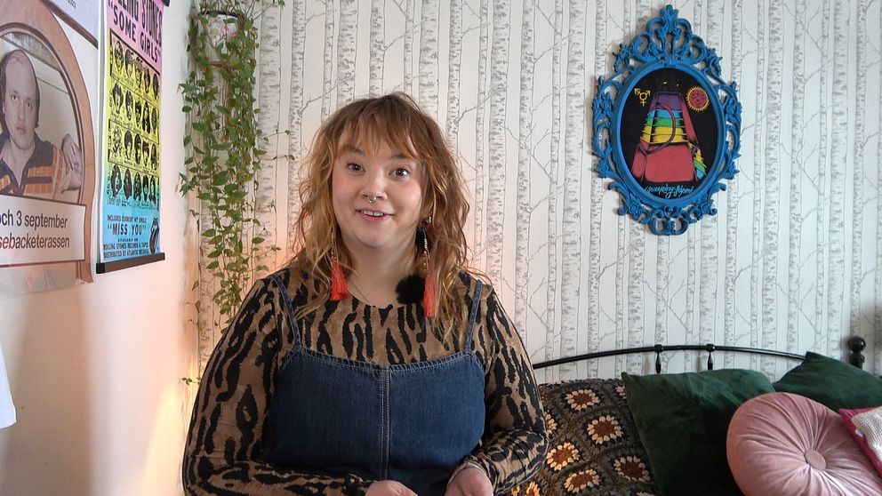 Person står i rum med väggdekoration och växter, iklädd en mönstrad tröja.