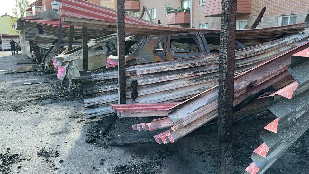 Skadade bilar parkerade under en delvis förstörd carport, med brända och lutande takplåtar. Markytan visar tydliga tecken på brand och förstörelse. I bakgrunden syns en byggnad med lägenheter.