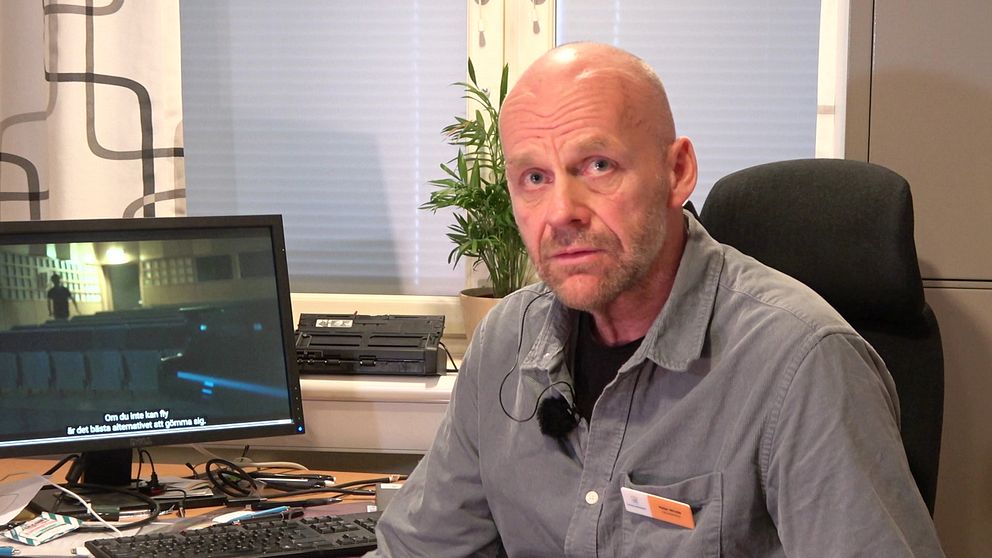 En person sitter vid ett skrivbord med en datorskärm i bakgrunden. Skärmen visar en scen från med någon som går i en skolaula. Ett skrivbord är belamrat med papper och kontorsutrustning, och det finns en grön växt i fönstret bakom.