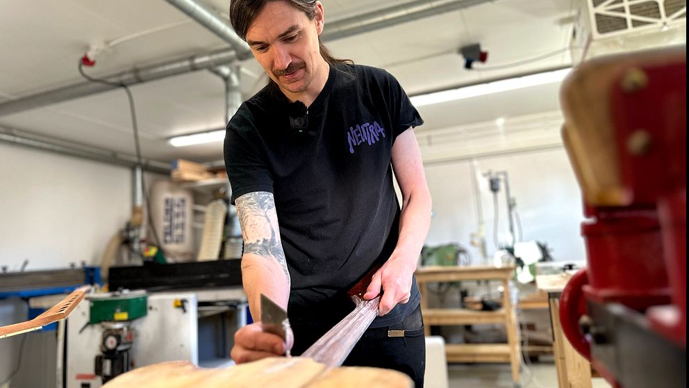 En person arbetar med en gitarr i en verkstad, använder ett verktyg för att forma eller slipa träet. Området är utrustat med olika verktyg och maskiner som syns i bakgrunden. Personens skjorta har en tryckt design.