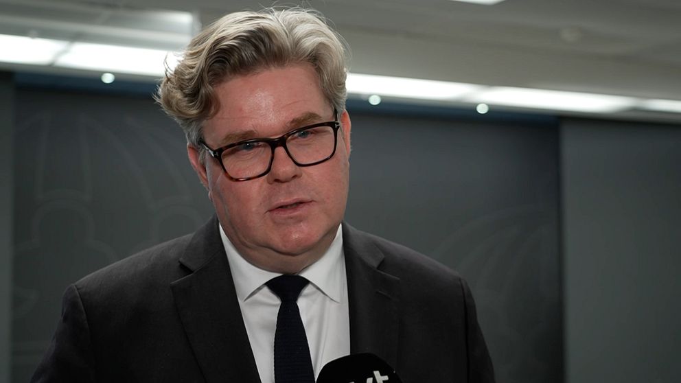 En person i kostym står framför en vägg med en subtil design, han ser ut att pratat i en mikrofon.