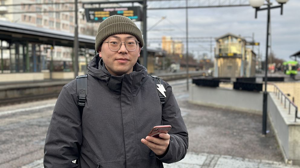 Person i mörk jacka står på en tågperrong och håller en smartphone.