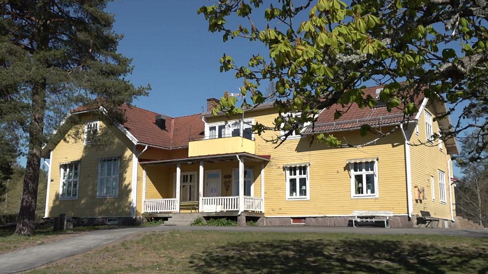 Gul trävilla med rött tak och veranda, omgiven av träd under klarblå himmel.